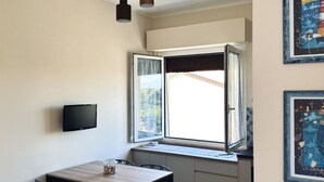 A room with a window, a table, chairs, a flat-screen TV, and artwork on the walls. - S340 - Sirolo, new two-room apartment in the center with parking space (Sirolo)