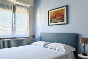 Un dormitorio con cama, un radiador, una ventana con persianas, un cuadro colgado en la pared y una lámpara de noche.