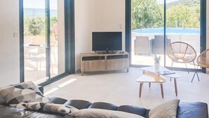 Ein modernes Wohnzimmer mit einer Ledersofa, einem Fernseher auf einem Holzkasten und Blick auf einen Swimmingpool durch große Fenster.