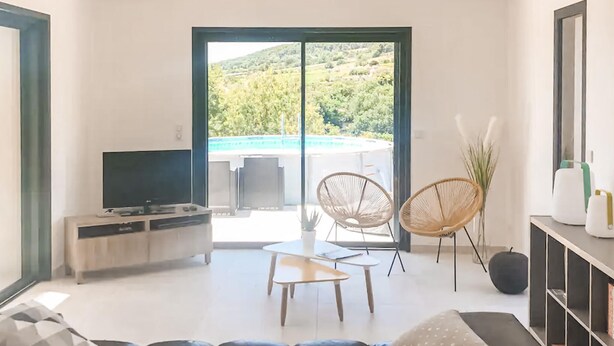 Ein modernes Wohnzimmer mit Sofa, Fernseher, kleinem Tisch, zwei Korbstühlen und Blick auf Pool und Grün hinter einer Glastür.