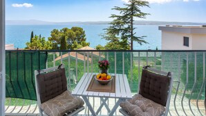 Ein Balkon mit zwei gepolsterten Stühlen, einem kleinen Tisch mit Obst und Blumenschale und Blick auf das Meer und die entfernten Hügel.