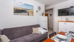 Ein modernes Wohnzimmer mit einem grauen Sofa, einem Esstisch gedeckt mit Tellern und Besteck, einem flachen Bildschirmfernseher über dem Tisch und einem Kühlschrank im Hintergrund.