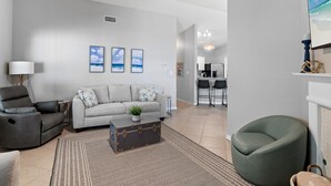 A modern living room with a grey sofa, a dark armchair, and a green bean bag chair. There is a coffee table with a vintage suitcase, a lamp, and a framed picture on the wall.