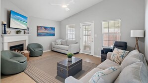 A spacious living room with a fireplace, a large flat-screen TV, a ceiling fan, and a view of the outdoors through the windows.
