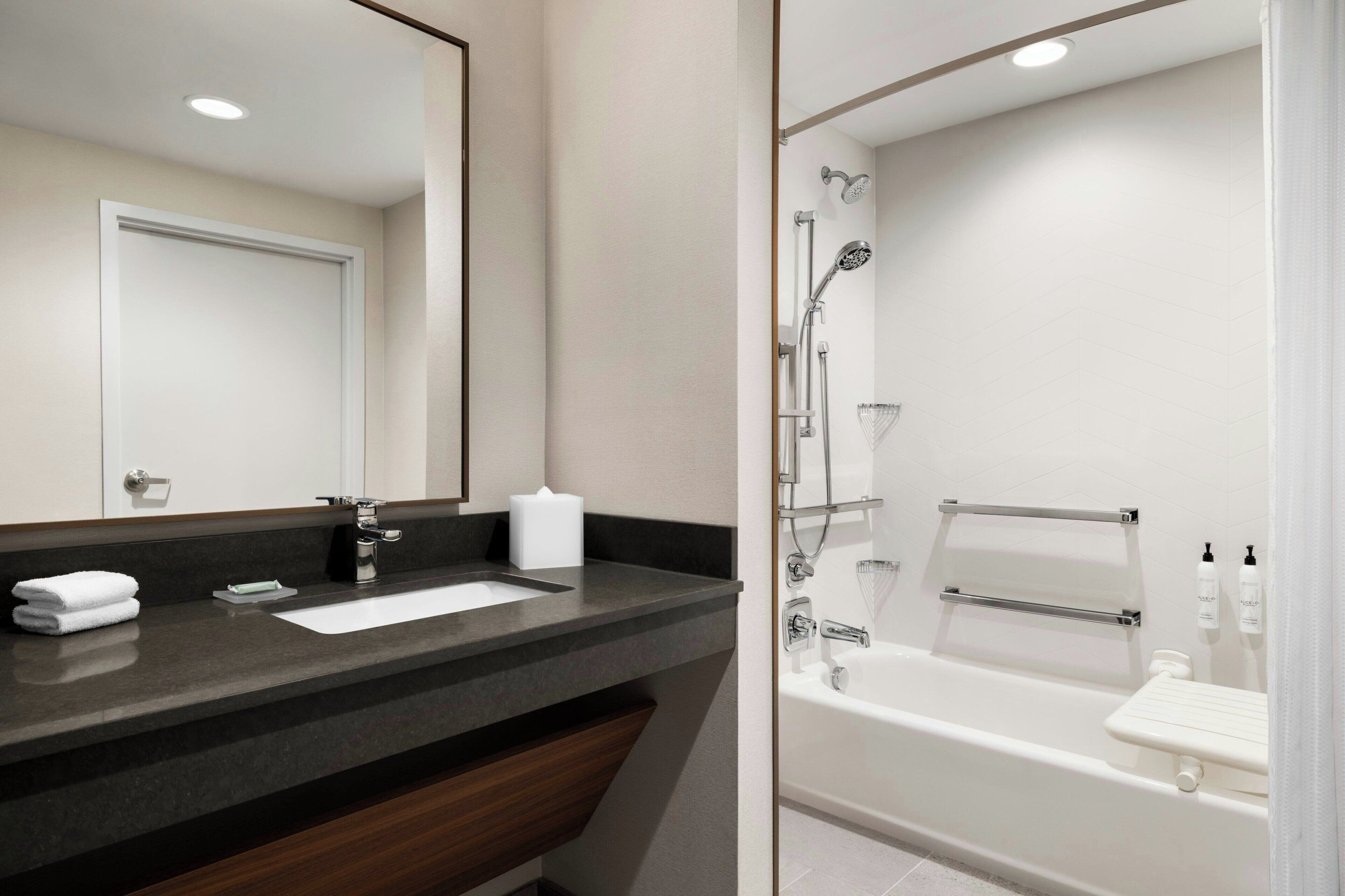 A modern bathroom with a dark countertop, a white sink, a large mirror, and a white bathtub with a handheld showerhead.
