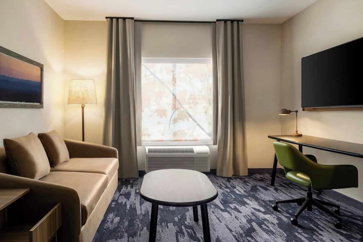 A hotel room with a sofa, a small table, a chair, a TV, and a window with curtains.