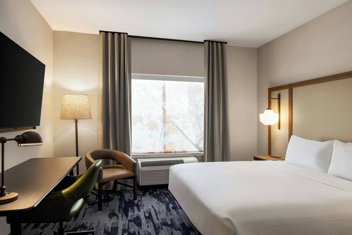 A hotel room with a bed, a desk with a chair, a TV, a lamp, and a window with curtains.