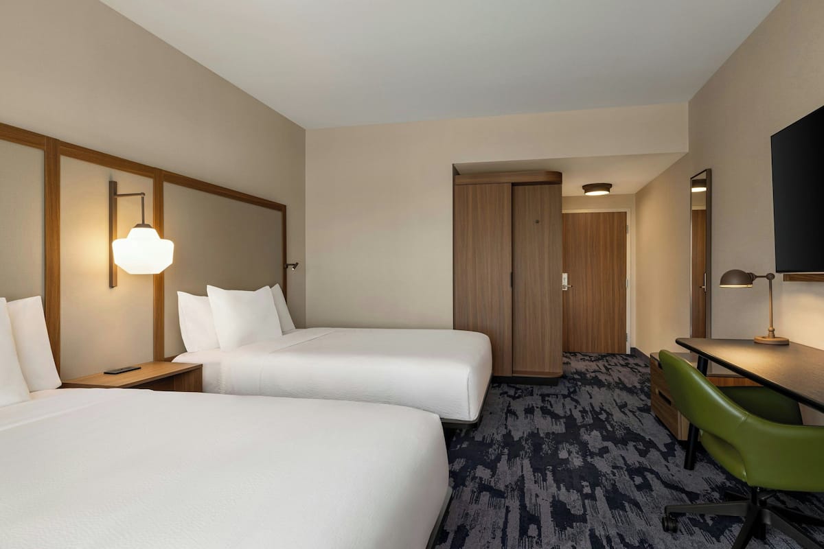 A hotel room with two beds, a desk, a green chair, a wooden wardrobe, and a wall-mounted TV.