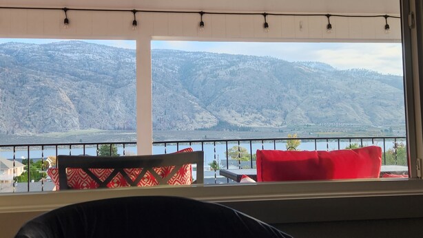 A room with a view of a lake and mountains. There is a window with a view of the lake and mountains. There is a chair with a red cushion and a black cushion. There is a railing in front of the window. - The View on 87th Osoyoos 2 bedroom, 1.5 bath, covered patio view vacation home. (Osoyoos)