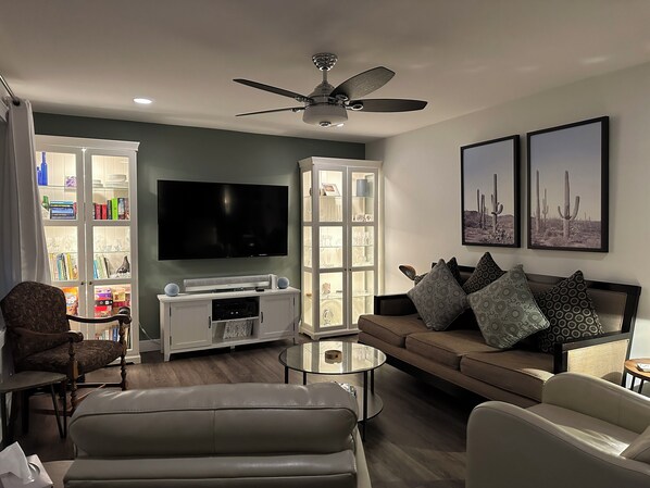 A living room with a flat-screen TV, a ceiling fan, a bookshelf, and a sofa with patterned cushions. - The View on 87th Osoyoos 2 bedroom, 1.5 bath, covered patio view vacation home. (Osoyoos)