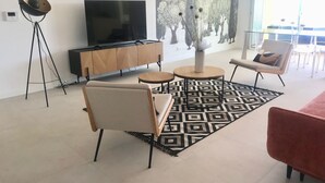 Ett modernt vardagsrum med en platt-TV, ett träskåp, en golvlampa, två fåtöljer, ett soffbord och en mönstrad matta.