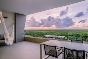 Uma varanda com mesa e cadeiras, uma rede e vista para uma paisagem verde com nuvens no céu.