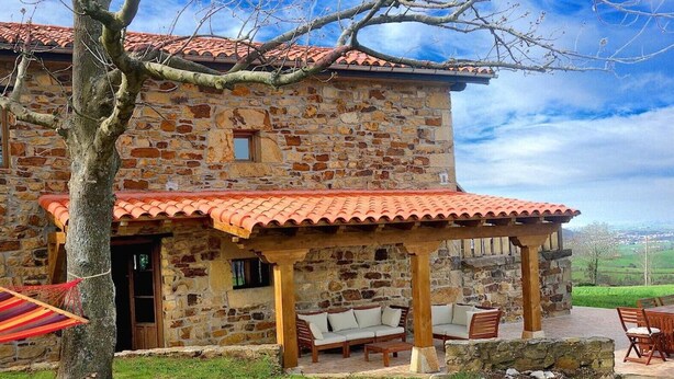 A stone building with a tiled roof, outdoor seating, and a tree in the foreground. - Country House 'La Casa Del Chileno' with Sea View, Private Terrace and Private Garden (Lierganes)