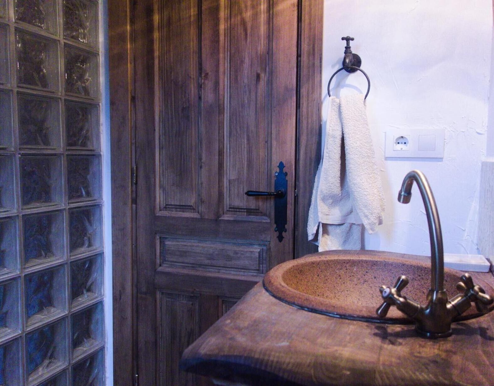 Un baño con puerta de madera, ventana de bloques de vidrio, una toalla colgada en un aro metálico y un lavamanos de piedra con un grifo antiguo.