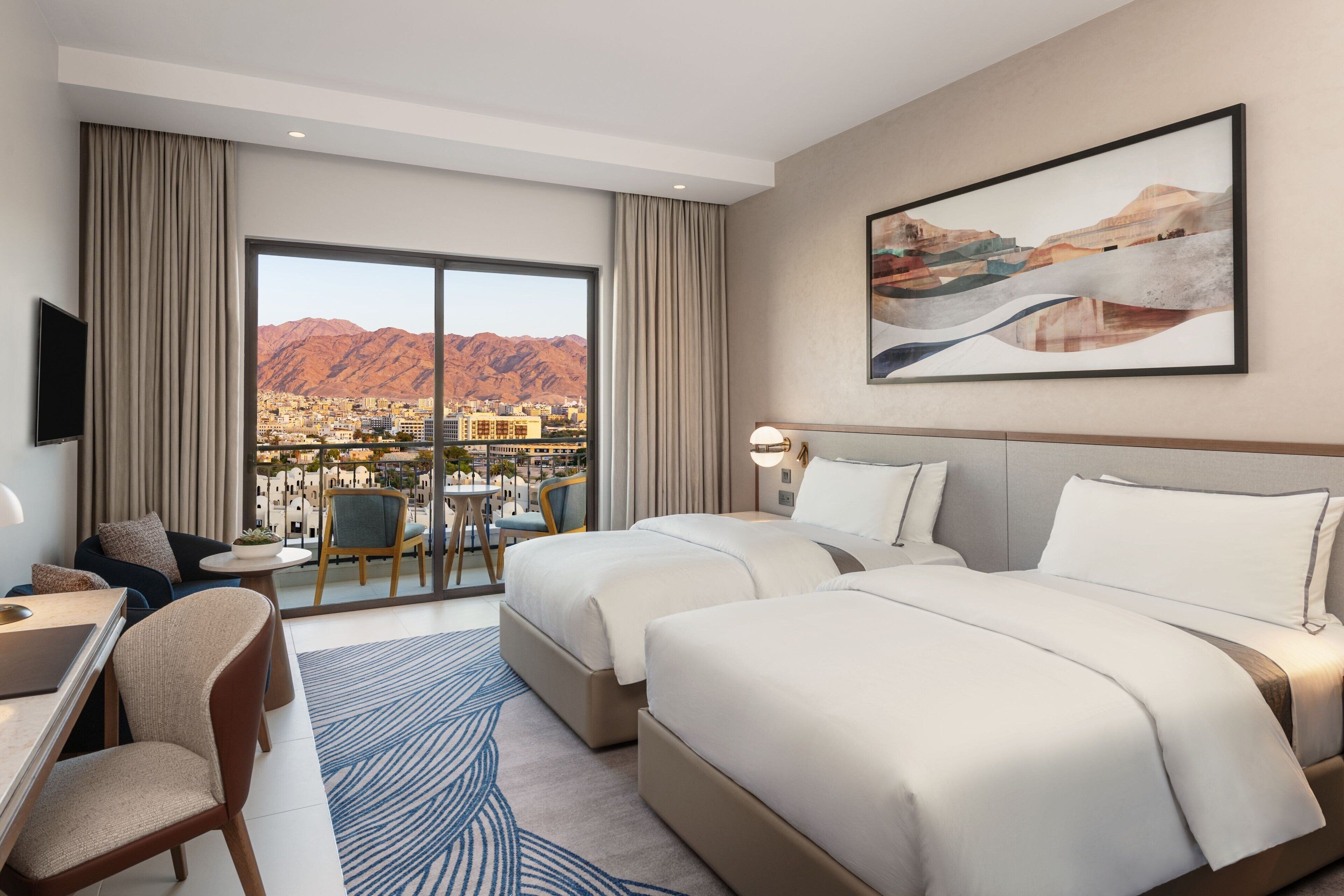 A hotel room with two beds, a desk, a chair, and a view of a city and mountains.