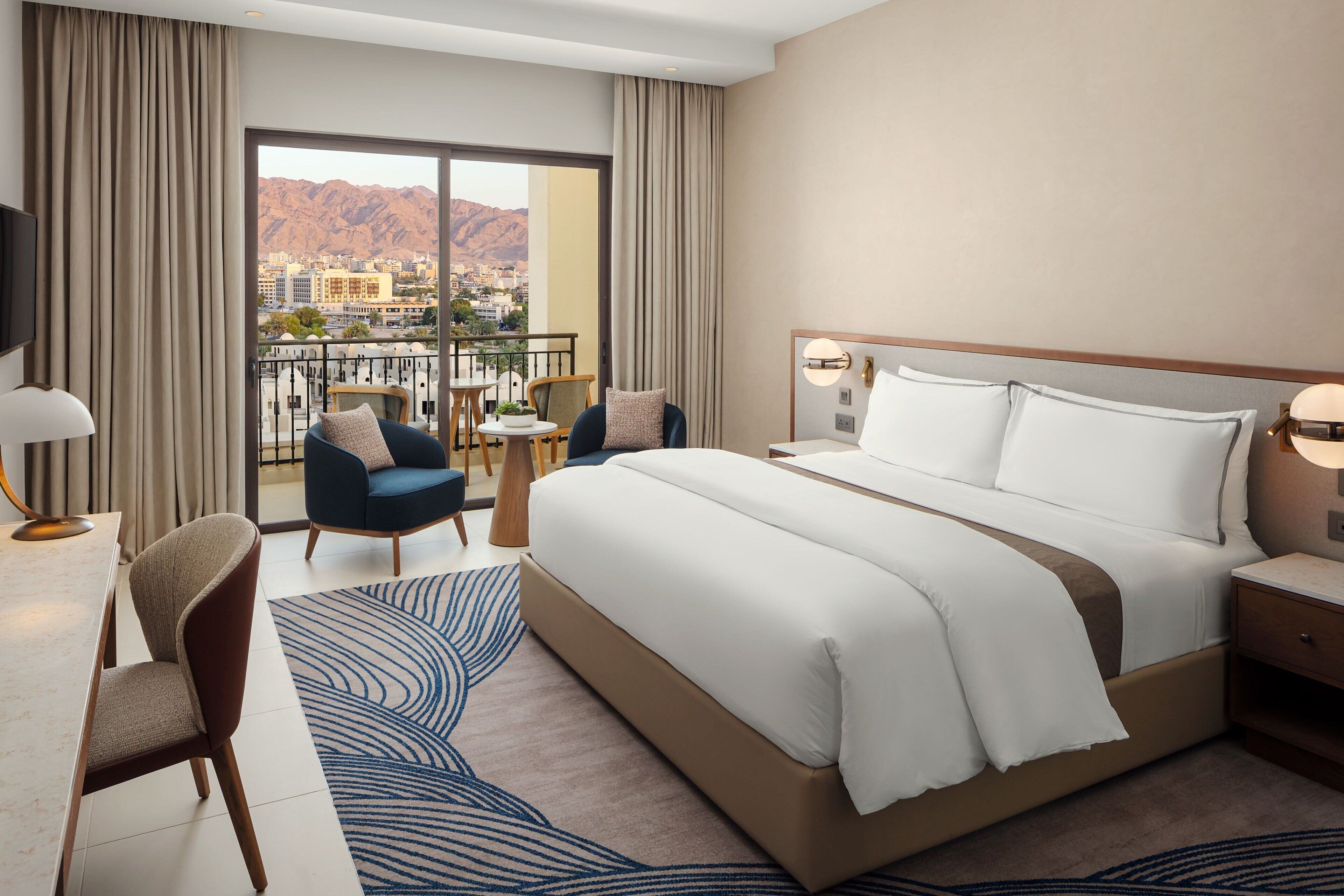 A hotel room with a large bed, a desk, a chair, a small table, and a balcony with a view of mountains.