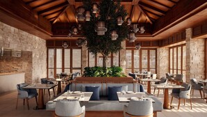Un restaurante espacioso con vigas de madera, paredes de piedra y lámparas colgantes. Las mesas están dispuestas con sillas, y hay un gran árbol en el centro.