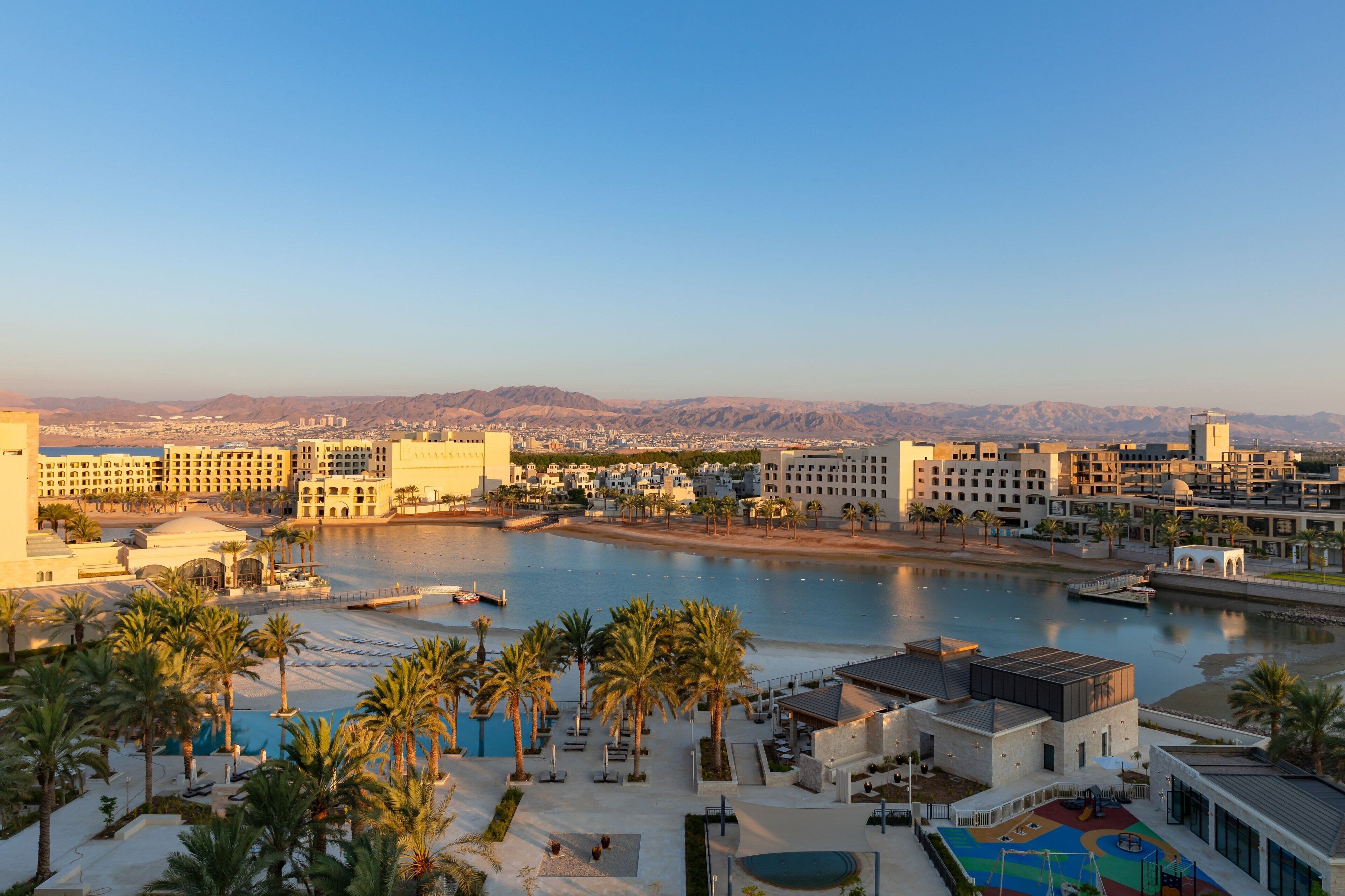 A hotel with a pool area, palm trees, and a view of a river and mountains.