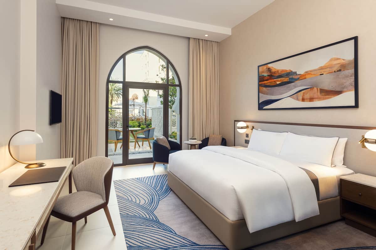 Habitación de hotel con una cama grande, un escritorio, una silla y vistas al exterior a través de una puerta de cristal.