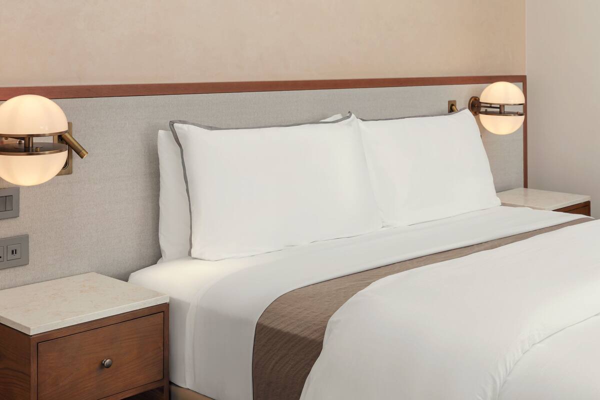 A neatly made bed with white linens and a brown accent, flanked by two bedside tables with lamps.