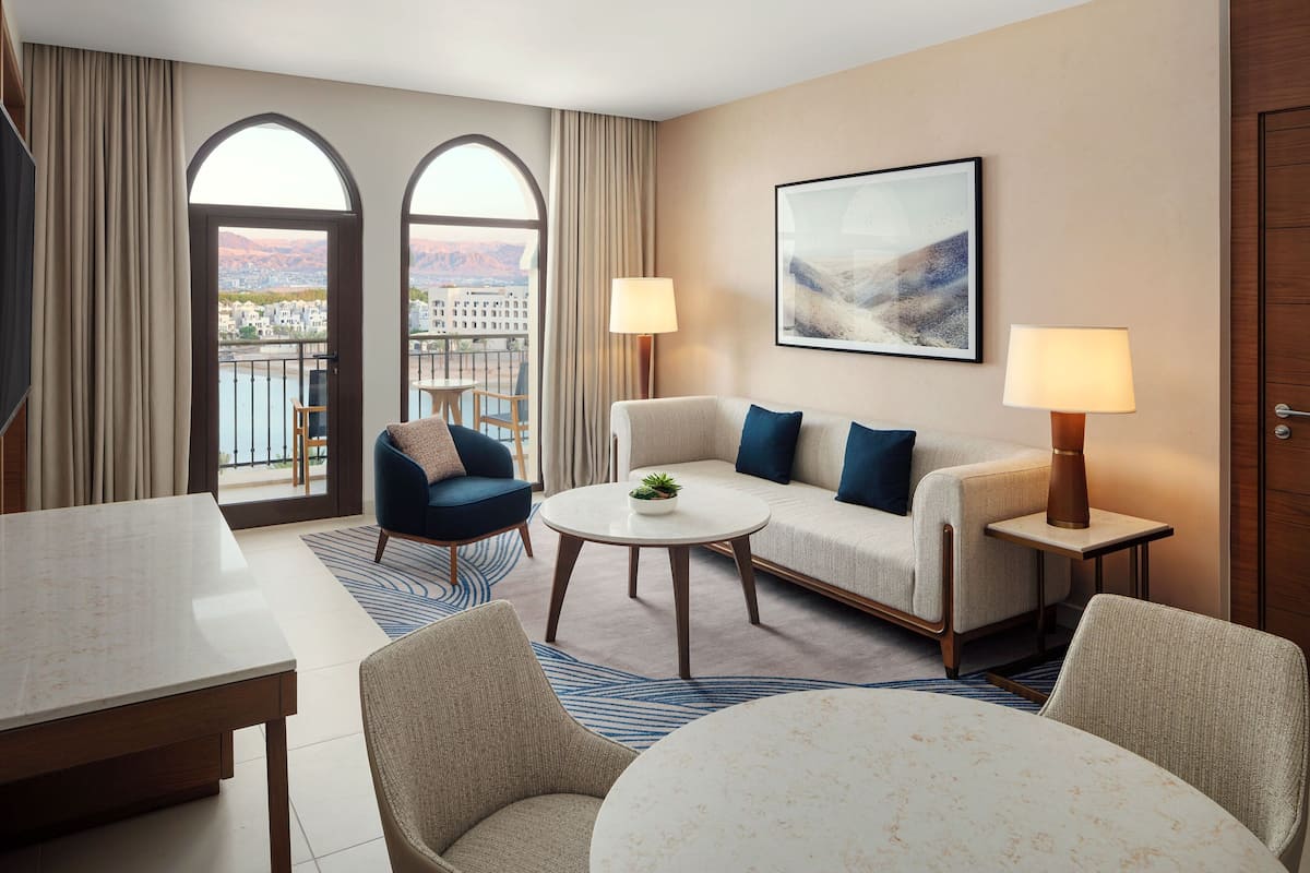 Una habitación de hotel con sofá, sillones, una mesa de comedor, una pequeña mesa redonda y vistas a un paisaje urbano a través de amplios ventanales.