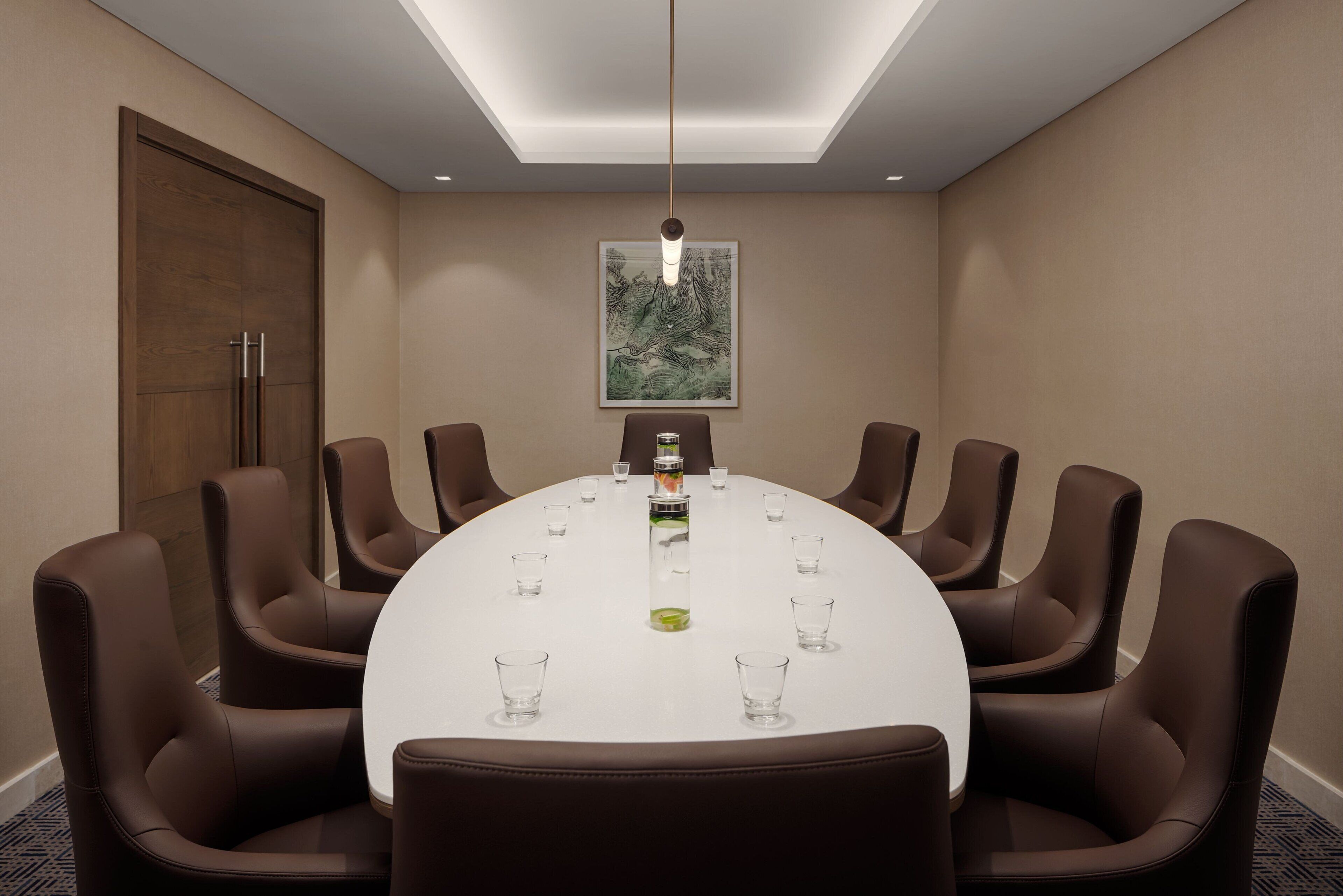 Un salón de conferencias con una mesa redonda, sillas de cuero marrón, un mantel blanco, una botella de agua en el centro y una obra de arte enmarcada en la pared.