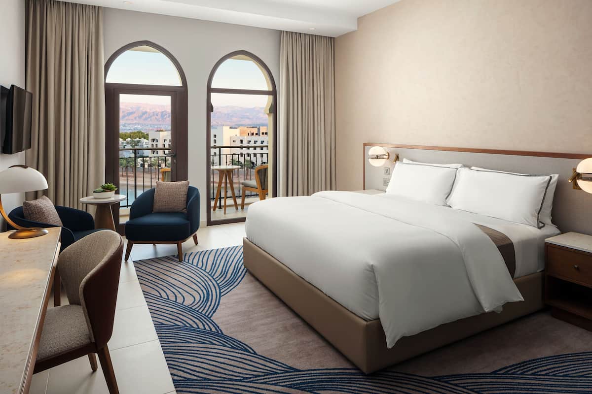 Habitación de hotel con una cama grande, un escritorio con silla, televisión y balcón con vistas a las montañas.