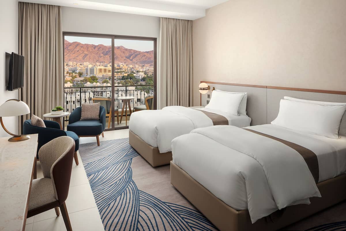Habitación de hotel con dos camas, un escritorio con silla, televisor y vistas a la ciudad y montañas.