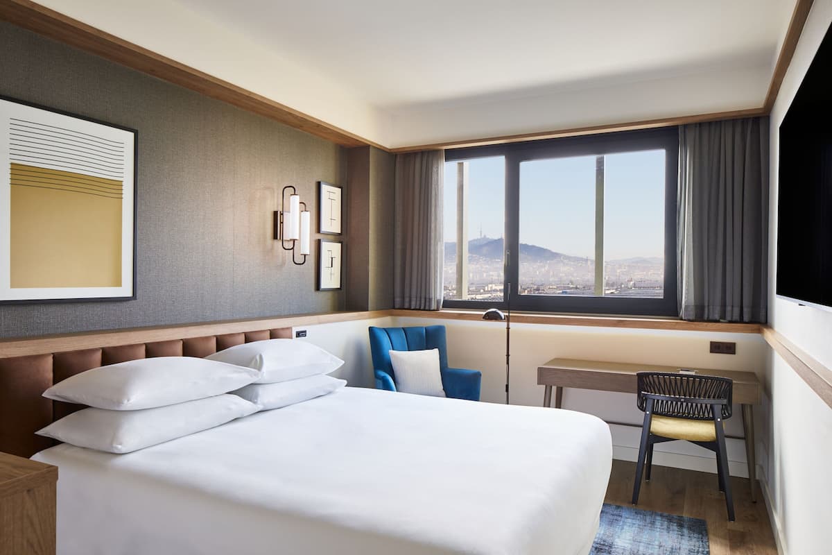 A hotel room with a bed, a desk, a chair, and a large window offering a city view.