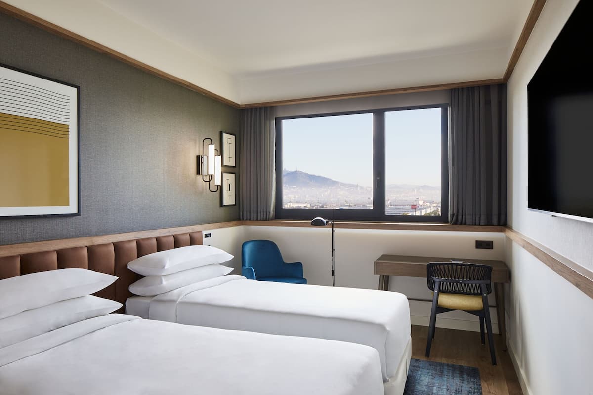 A hotel room with two beds, a desk, a chair, and a large window with a city view.