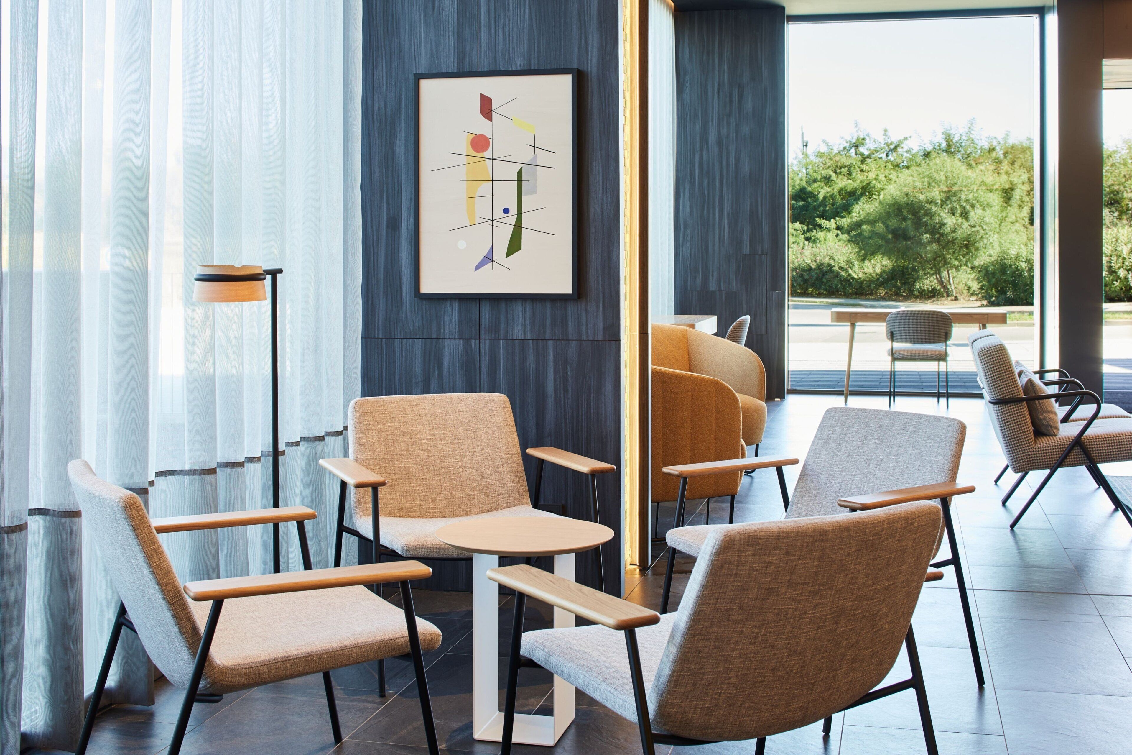 A modern interior with a seating area featuring chairs and tables, a framed abstract artwork on the wall, and a view of greenery outside.