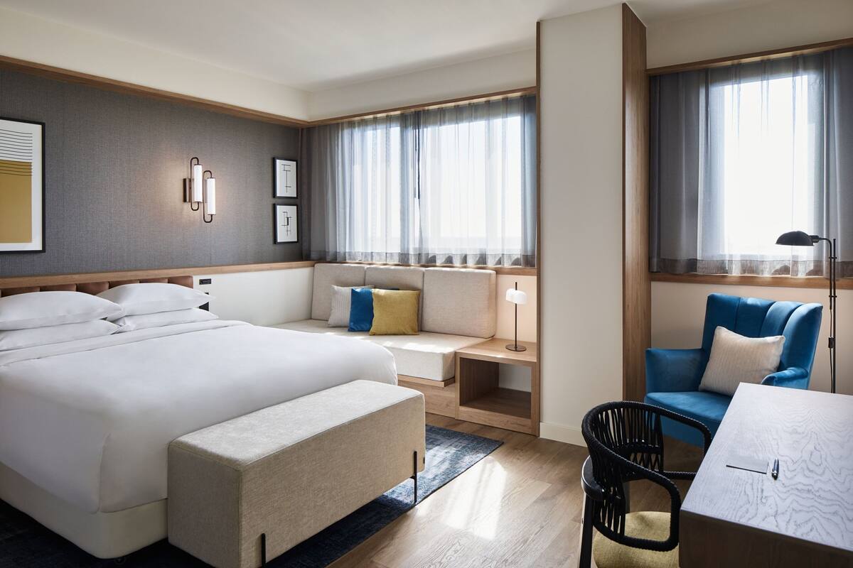 A hotel room with a large bed, a desk with a chair, a sofa, and a window with curtains.