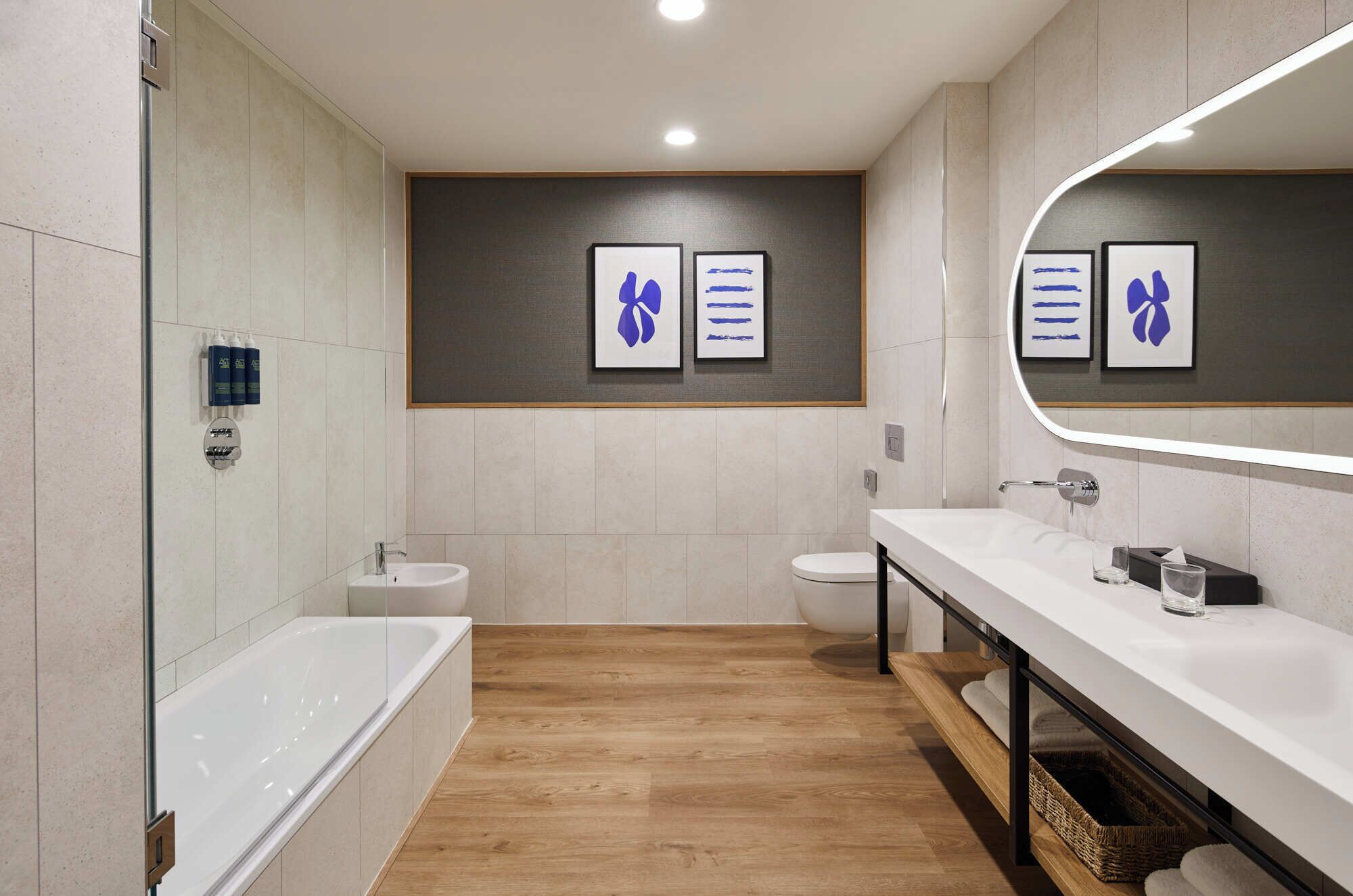 Een moderne badkamer met een badkuip, een wastafel en een toilet. Er hangen kunstwerken in lijsten aan de muur, en boven de wastafel hangt een grote spiegel.