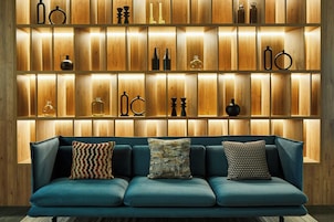 Uma sala de estar moderna com um sofá azul e almofadas estampadas, uma parede de madeira com prateleiras exibindo diversos objetos decorativos.