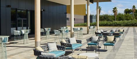 Area esterna con diversi tavoli e sedie, una zona coperta e un edificio con ampie finestre sullo sfondo.