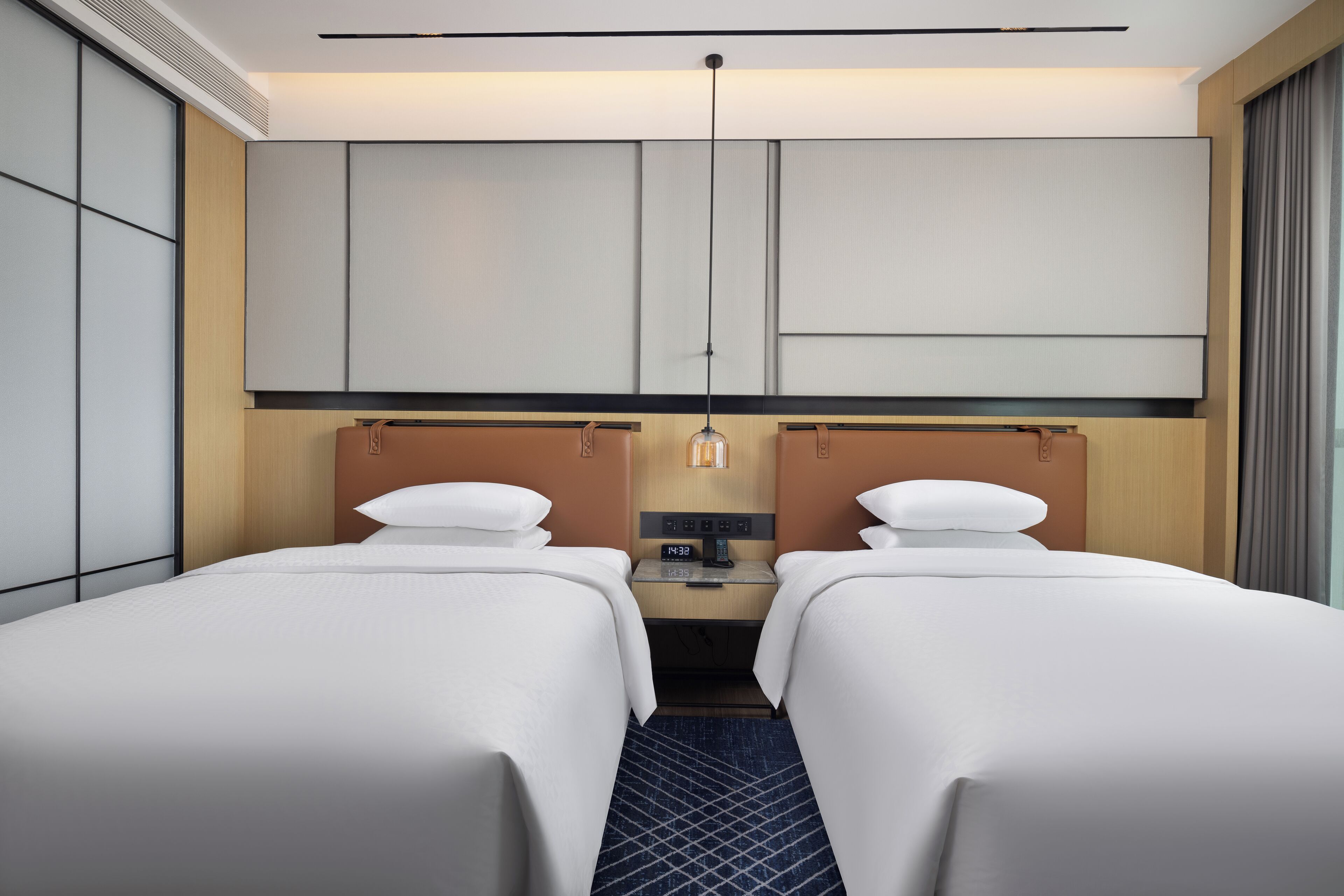 Een hotelkamer met twee bedden, een klein bureau, een nachtkastje en een groot raam met gordijnen.