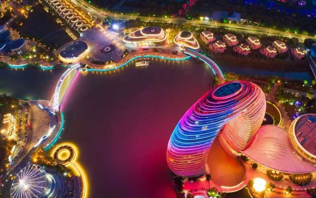 Un edificio grande y de diseño único, con forma circular, rodeado por una zona ribereña con senderos iluminados y una masa de agua.