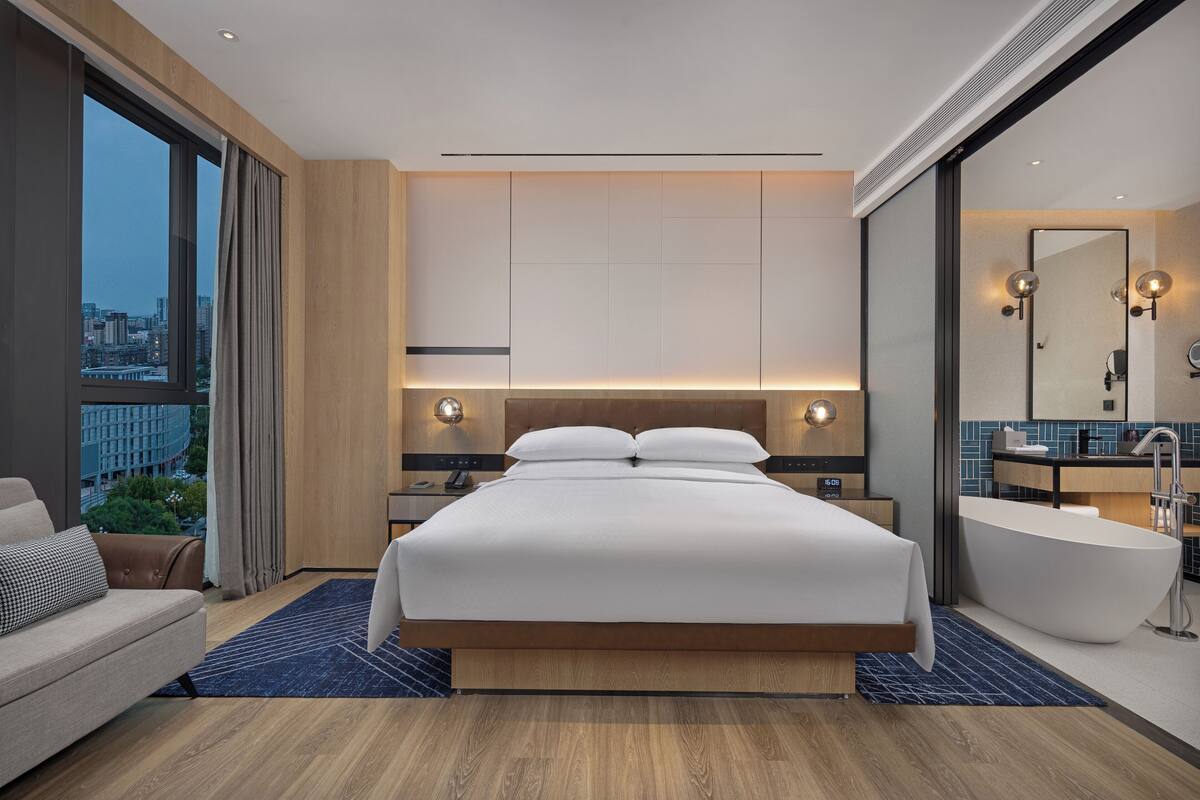 Une chambre d’hôtel moderne dotée d’un grand lit, d’un coin salon, d’une baignoire et offrant une vue sur la ville.