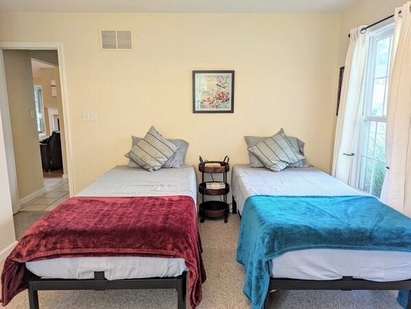 Una habitación con dos camas, cada una con una manta de color diferente, una mesita con una cesta y un cuadro en la pared.