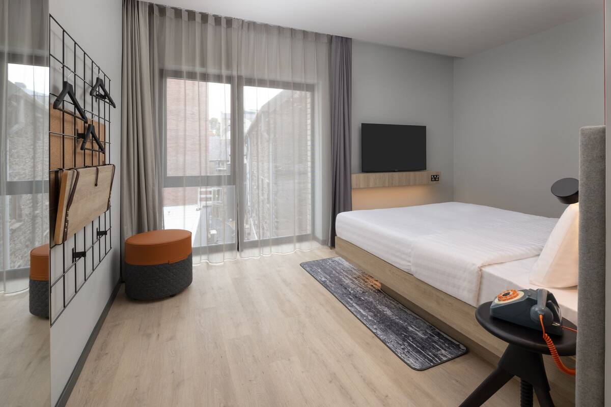 A modern hotel room with a bed, a TV, a desk with a telephone, a seating area with a chair, and a window with curtains.