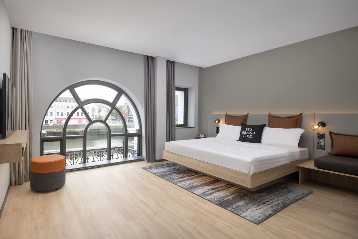A modern hotel room with a large bed, a view of a river, and a balcony with a railing.