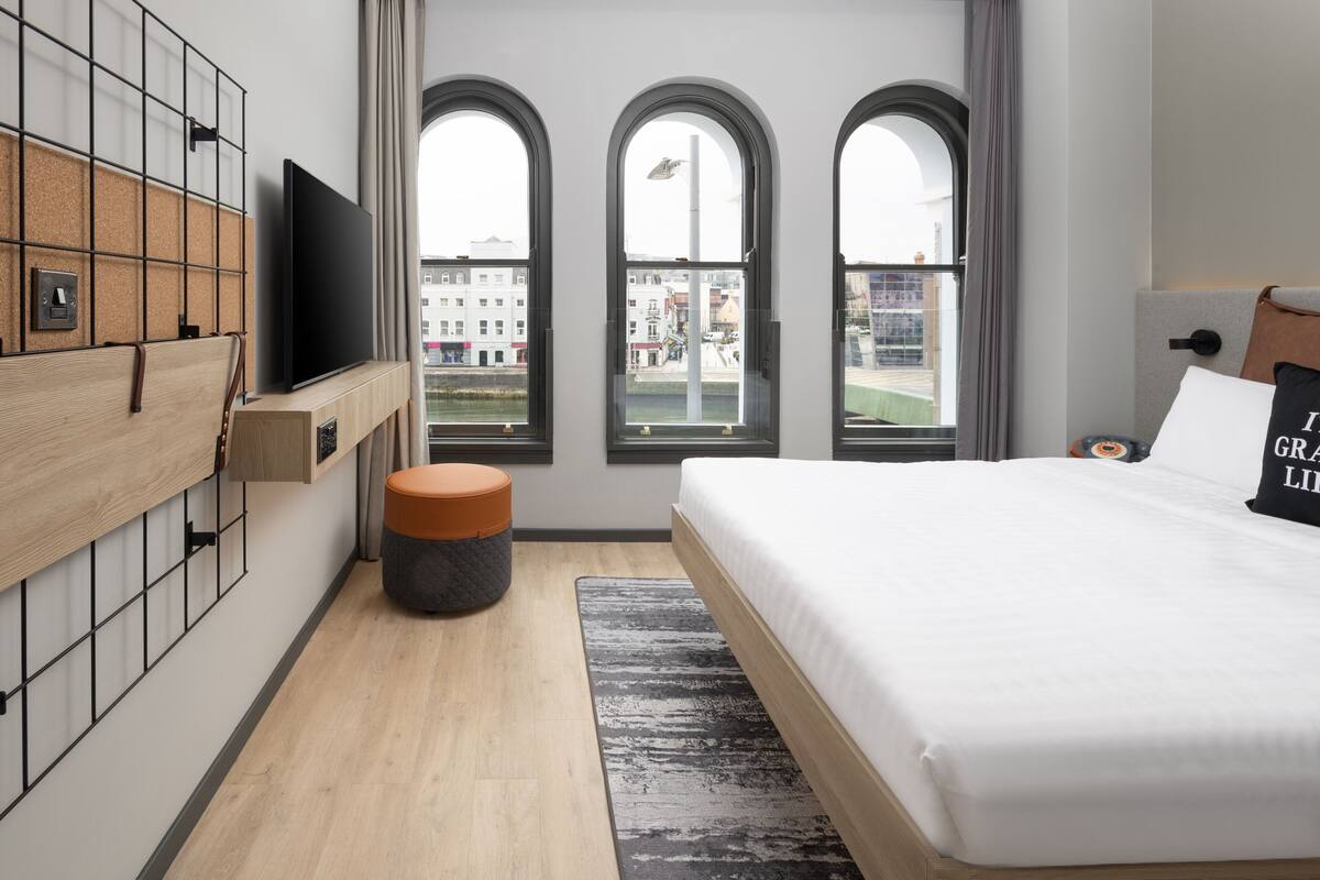 A modern bedroom with a large bed, a TV mounted on a wooden panel, and a view of the city through arched windows.