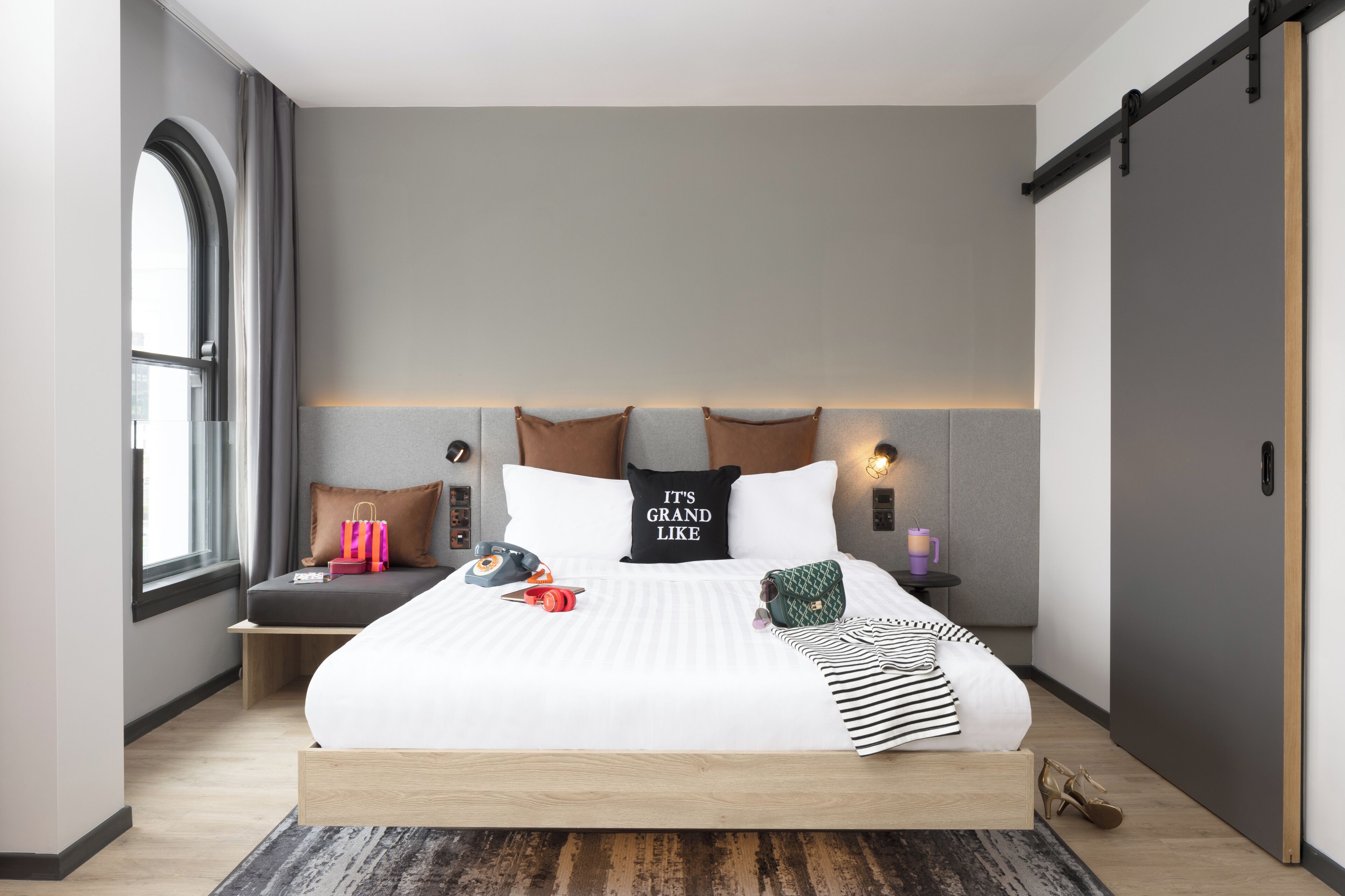 A modern bedroom with a large bed, a headboard with built-in lighting, a bedside table with a lamp, and a window with curtains.