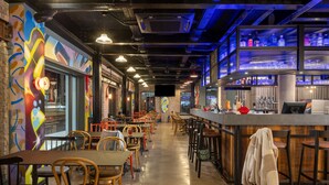 Zona de bar con mesas y sillas, una barra con caja registradora y estantes con botellas.