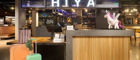 A hotel reception area with a large illuminated sign reading "HIYA" above the counter, a plush seating area, and a whimsical unicorn statue.