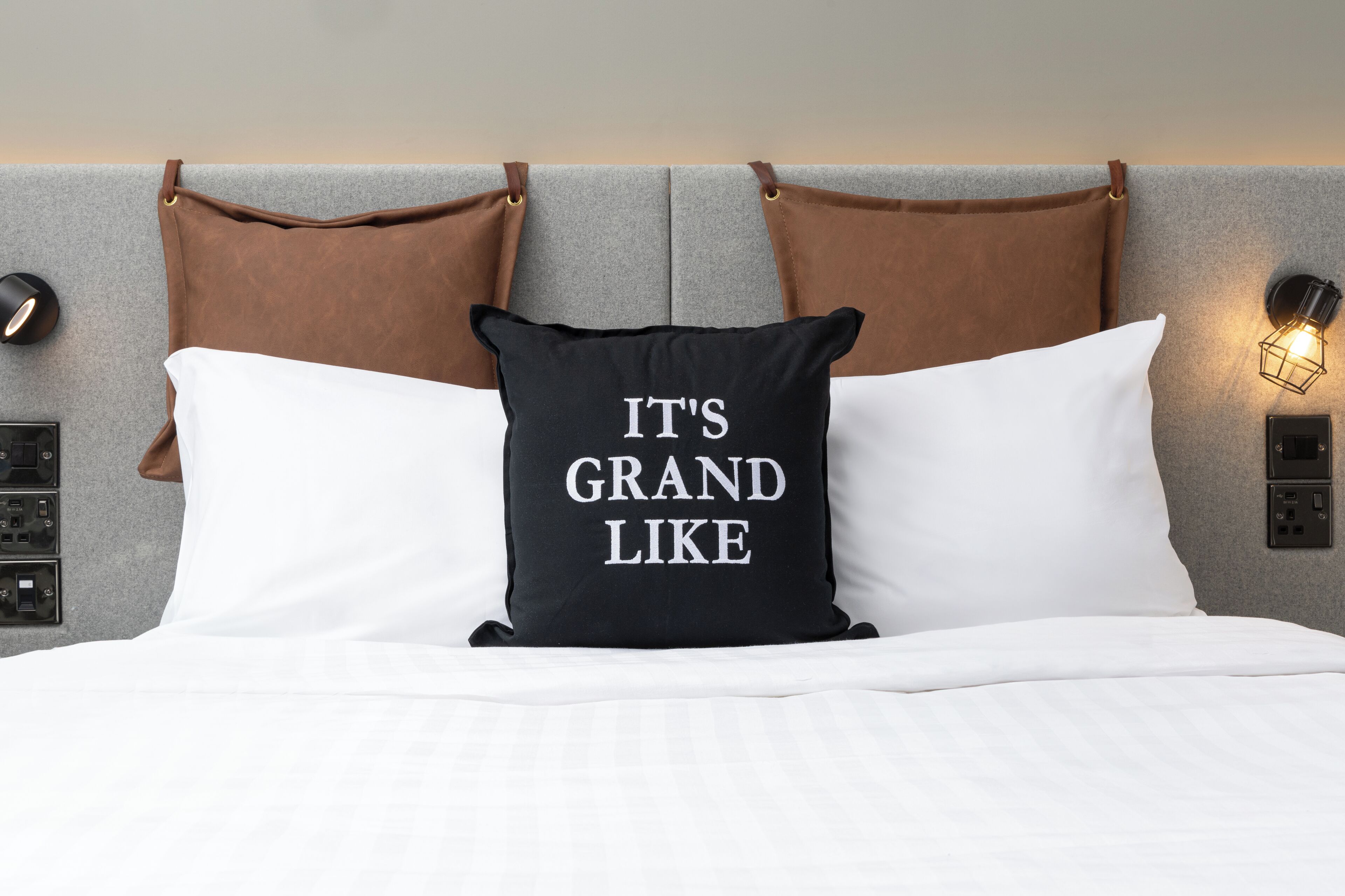A bed with white bedding, a black pillow with "IT'S GRAND LIKE" text, and brown leather headboard straps.
