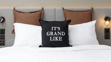 A bed with white bedding, a black pillow with "IT'S GRAND LIKE" text, and brown leather headboard straps.