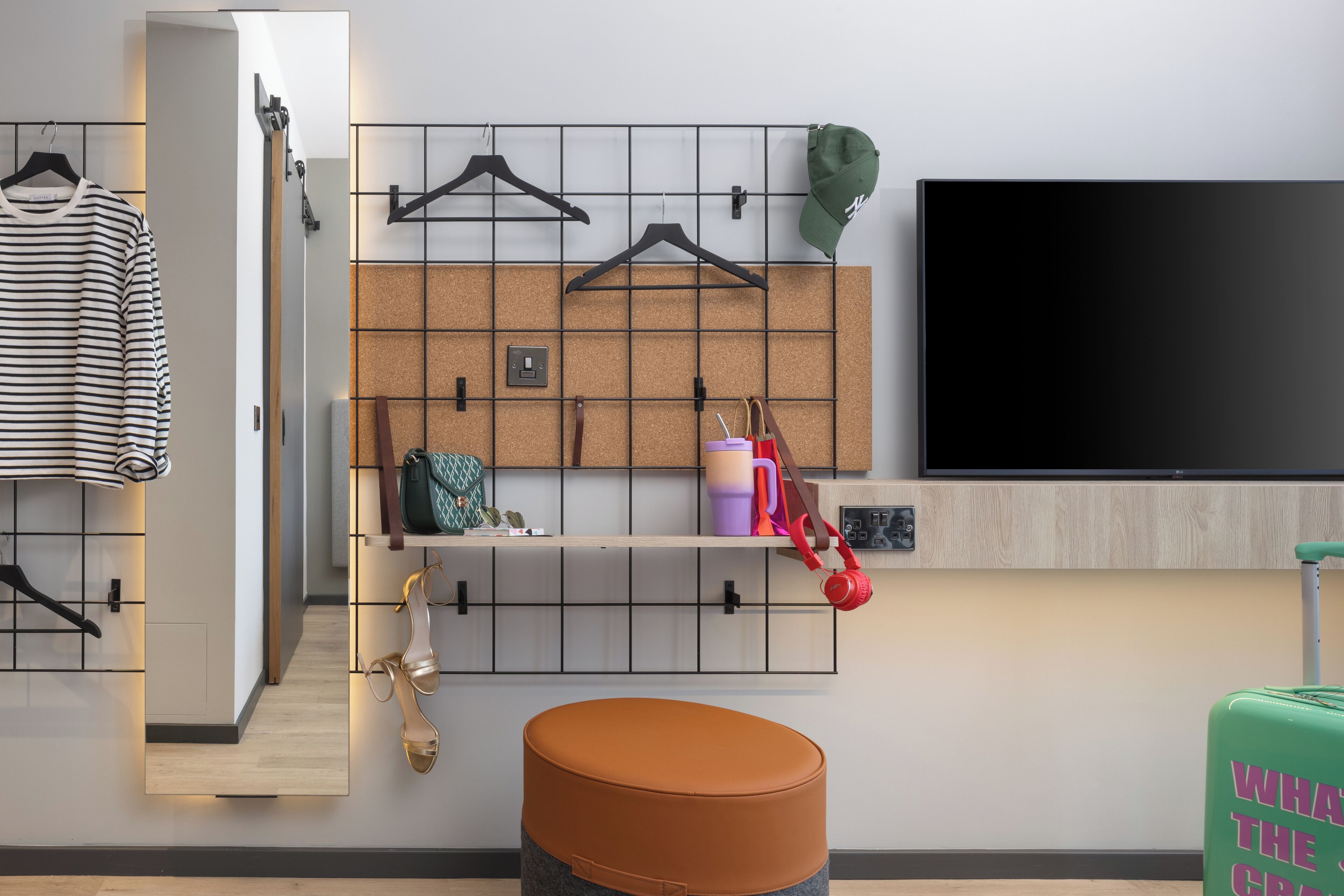 A modern hotel room with a corkboard wall, a flat-screen TV, a wooden bench, and a green suitcase.