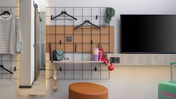 A modern hotel room with a corkboard wall, a flat-screen TV, a wooden bench, and a green suitcase.