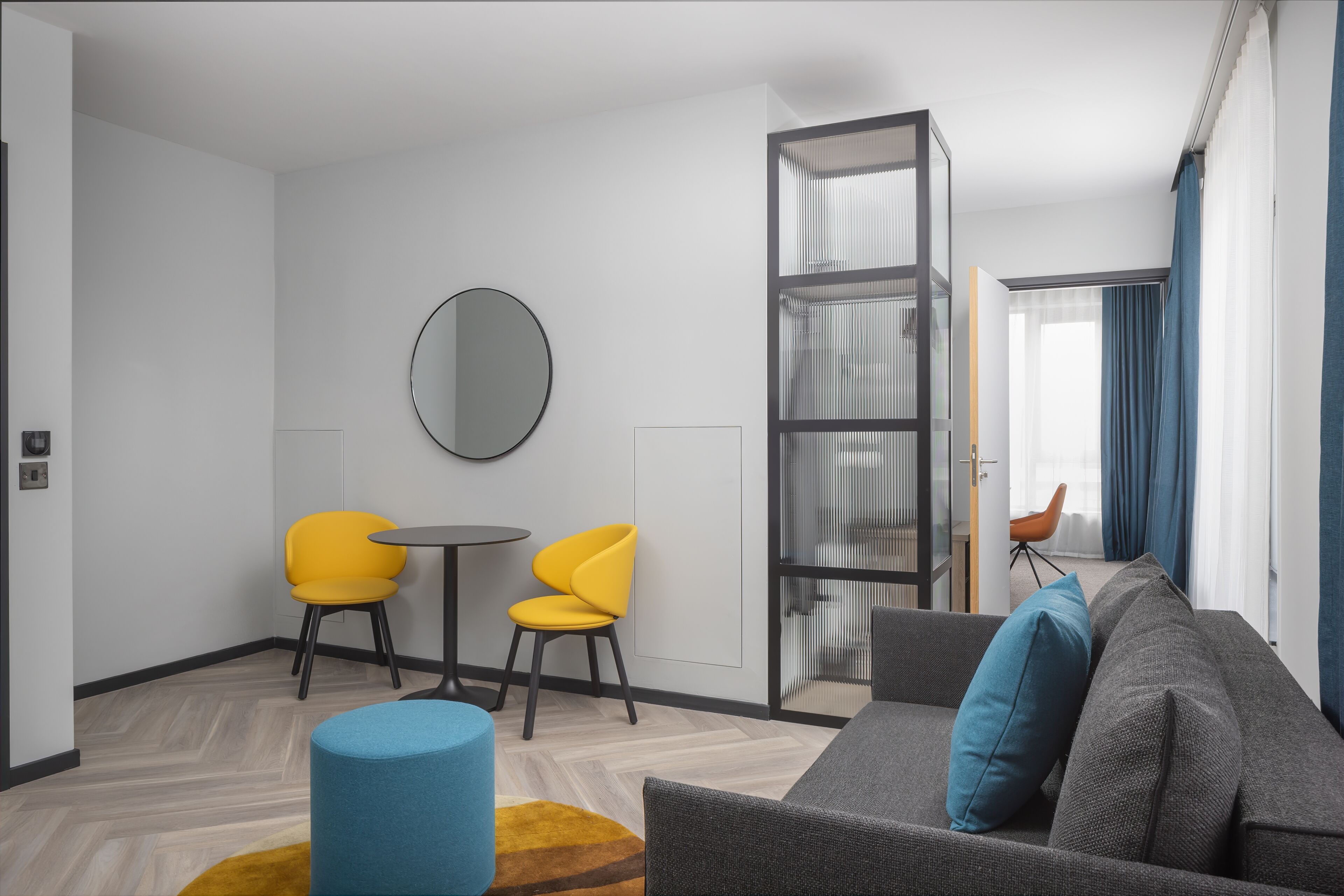 A modern living room with a sofa, two yellow chairs, a round mirror, and a glass partition.
